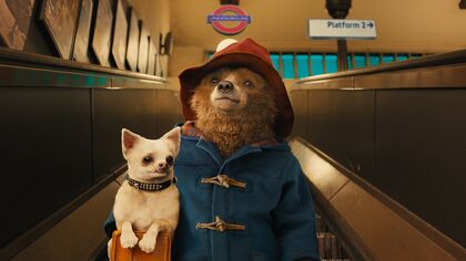 Paddington bear wearing a blue coat and red hat, carries a small dog and suitcase while riding down an escalator on the London underground